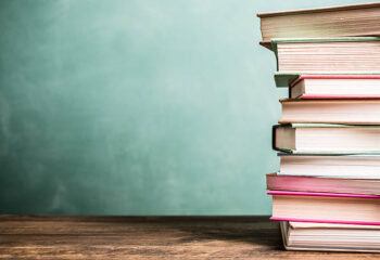 It's back to school time!  A large stack of textbooks to side makes frame composition.  The pile of objects lies on top of a wooden school desk with a green chalkboard in the background.  The blank blackboard in the background makes perfect copyspace!  Education background themes.